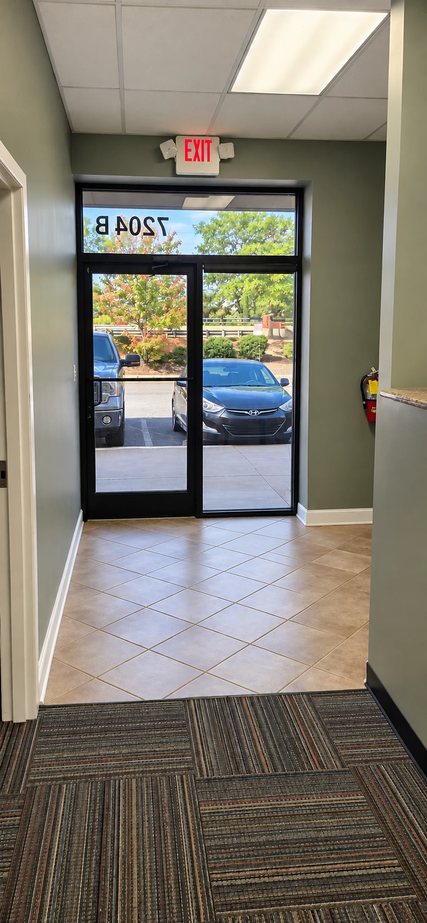 Suite B entry at 7204 Stinson Hartis Road: a tiled vestibule with sage-green walls leading to a glass storefront door marked 7204 B, with parking and landscaping outside
